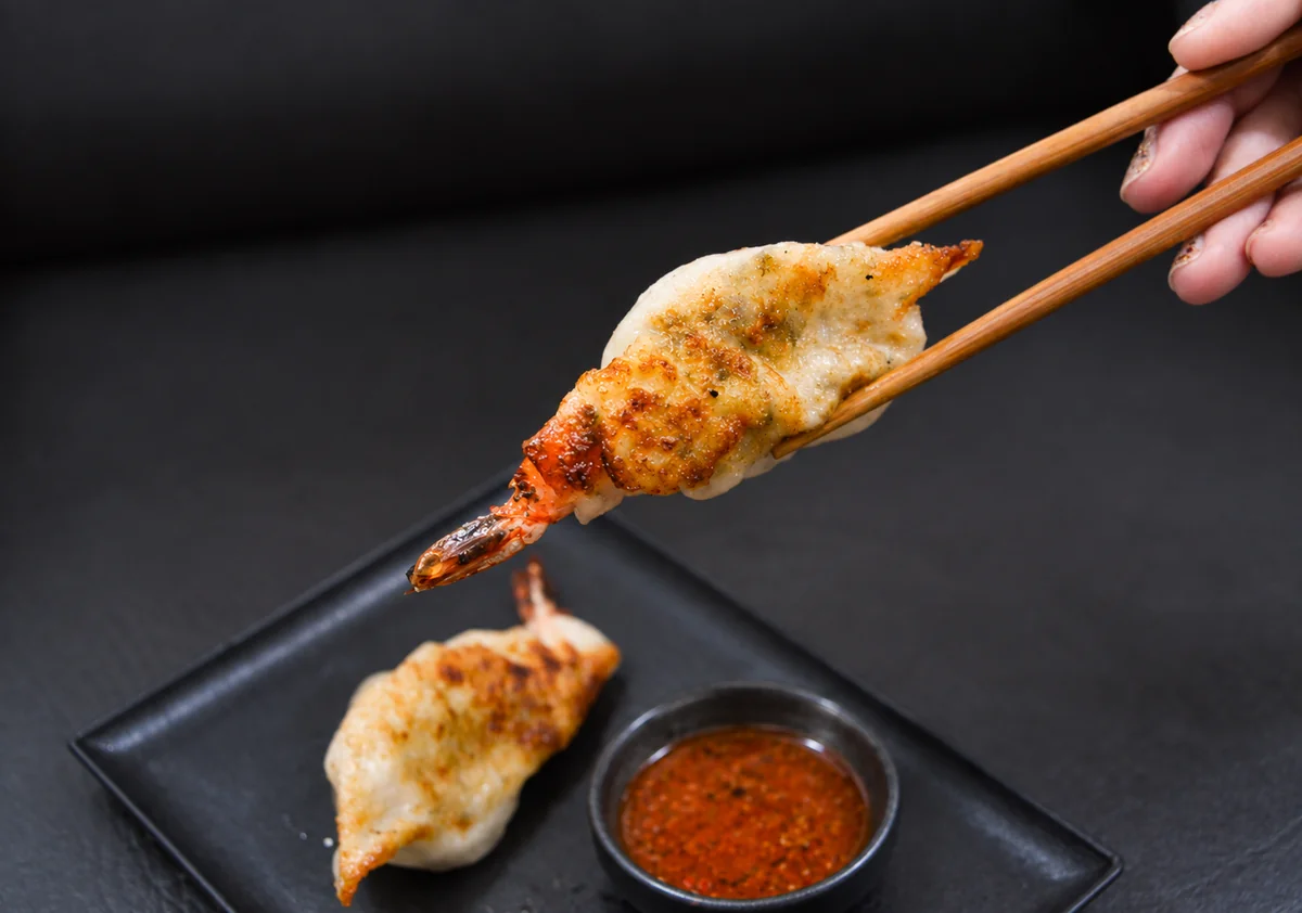 A shrimp Cajun gyoza held with wooden chopsticks above a black plate